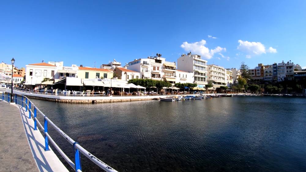 A picturesque waterfront view in Agios Nikolaos, Crete, featuring a calm bay, boats, and charming buildings under a clear blue sky. | SmartCarRental