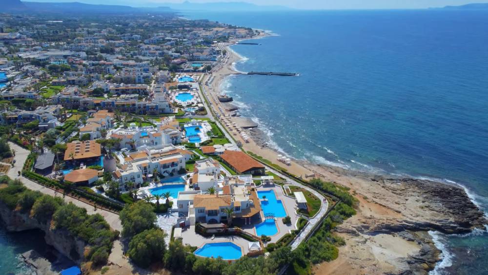 Vue aérienne d'Anissaras, en Crète, montrant une zone de villégiature côtière avec plusieurs piscines, des propriétés en bord de mer et la vaste mer bleue sous un ciel clair. | Cheap Car Rental