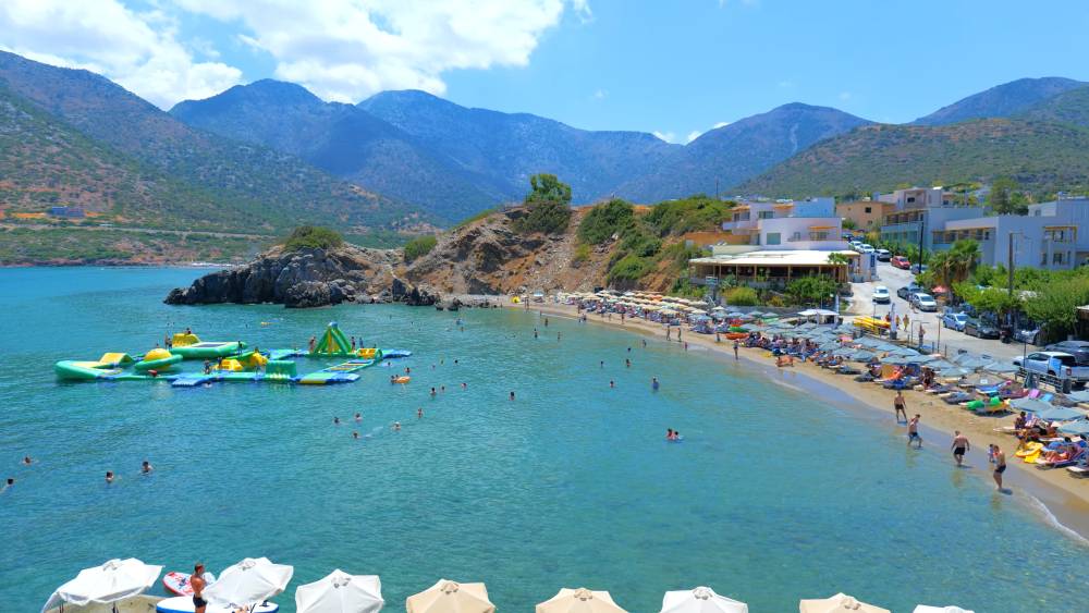 Beach view at Bali, Crete, featuring a scenic bay with calm turquoise waters, people enjoying water activities and sunbathing, and a backdrop of mountains and coastal buildings | Smart Car Rental