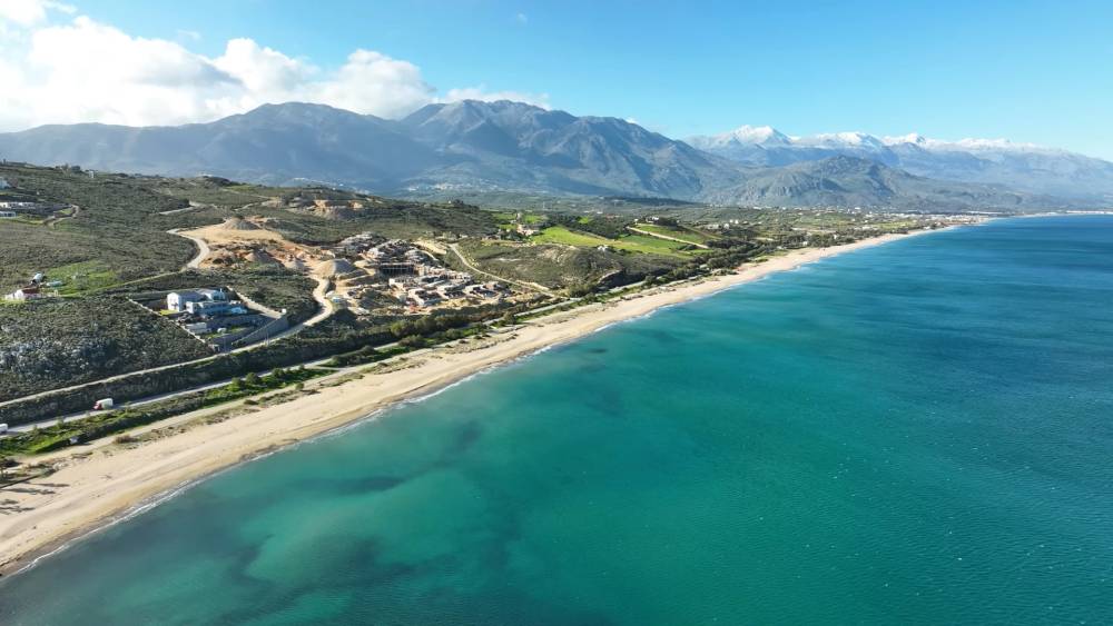 Aerial view of Episkopi Beach in Crete, showcasing the expansive coastline with clear blue waters, sandy shore, and mountainous landscape in the background. | Cheap Car Rental