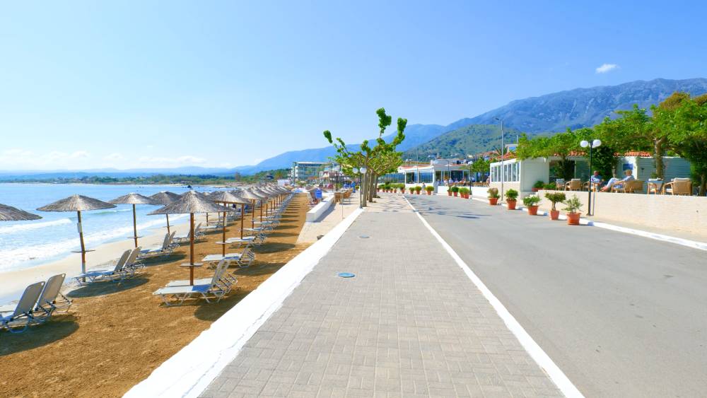 Vista panoramica della passeggiata sul lungomare di Georgioupolis, Creta, con file di lettini e ombrelloni lungo la spiaggia sabbiosa e montagne sullo sfondo | Smart Car Rental