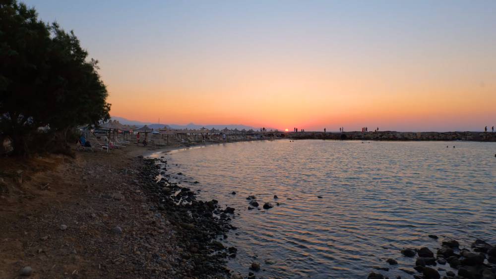 Sonnenuntergang in Gouves, Kreta, mit einem ruhigen Strand mit Sonnenliegen, einer felsigen Küste und Silhouetten von Menschen, die die ruhige Meeresatmosphäre genießen. | Smart Car Rental