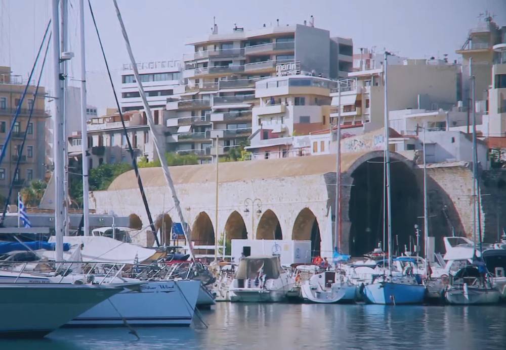 Vue des bateaux amarrés au port d'Héraklion en Crète, avec l'arsenal vénitien et les bâtiments modernes en arrière-plan, montrant un mélange d'architecture historique et contemporaine. | Cheap Car Rental
