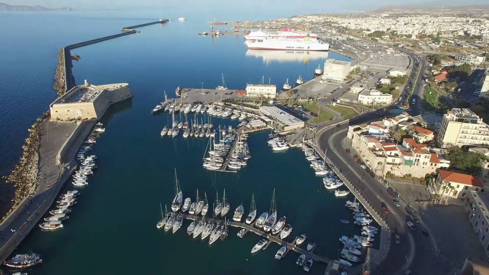 Vue aérienne du port d'Héraklion en Crète, avec de nombreux bateaux amarrés dans la marina, la forteresse vénitienne et un grand ferry en arrière-plan, avec la ville d'Héraklion entourant le port. | Smart Car Rental