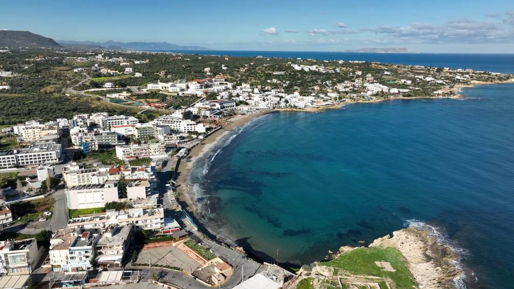 Aerial view of Hersonissos, showing the coastline, buildings, and clear blue waters from a drone perspective. | Cheap car rental