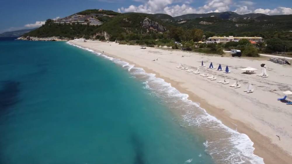 Breiter Strandblick bei Karavostasi, Kreta, mit einem unberührten Sandstrand mit türkisfarbenem Wasser, Reihen von Strandstühlen und Sonnenschirmen, und einem Hintergrund aus üppigen grünen Hügeln und Gebäuden | Günstige Autovermietung