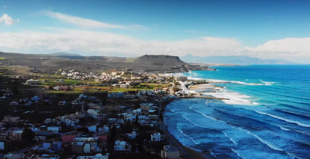 Aerial view of Kokkini Hani, Crete, featuring a coastal town with white buildings, rolling waves along the shoreline, and a mountainous landscape under a bright blue sky. | Cheap Car Rental