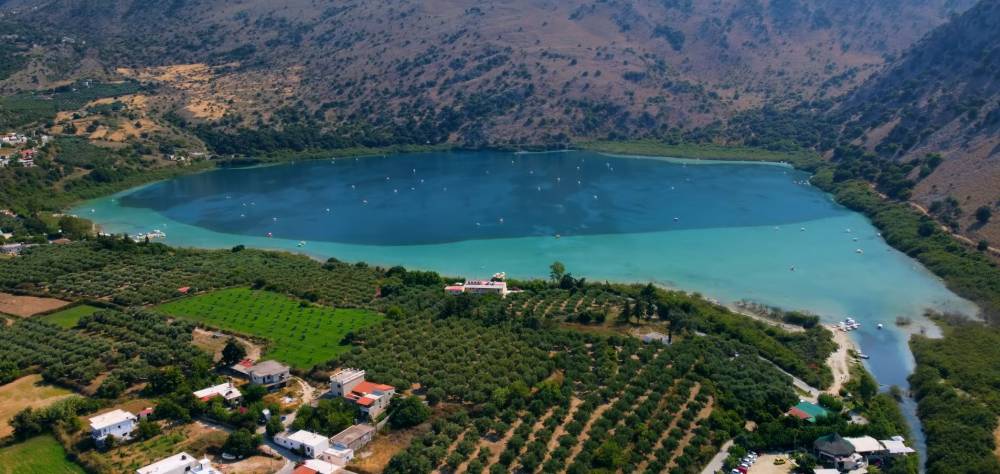 Aerial view of Lake Kournas in Crete, surrounded by lush greenery and hills, with clear turquoise waters and small boats on the lake | Cheap Car Rental