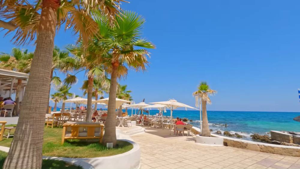 A beautiful seaside restaurant in Malia, Crete, Greece, featuring palm trees, outdoor dining, and clear blue waters of the Mediterranean Sea. | Cheap Car Rental