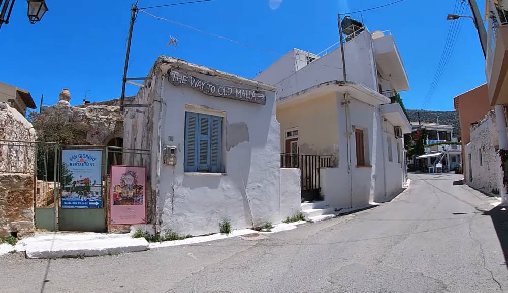 Vue de rue de la vieille ville historique de Malia, en Crète, avec un panneau indiquant 'The way to Old Malia' et des bâtiments traditionnels en arrière-plan | Cheap Car Rental