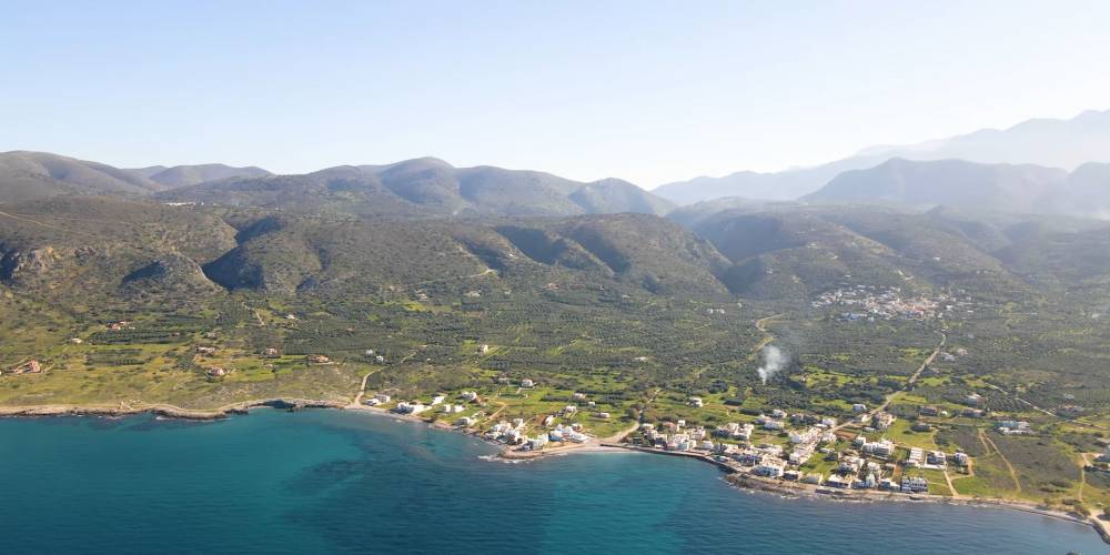 An aerial view of Milatos, a coastal village in Crete, Greece, featuring green hills, mountains, and the clear blue waters of the Mediterranean Sea. | Smart Car Rental