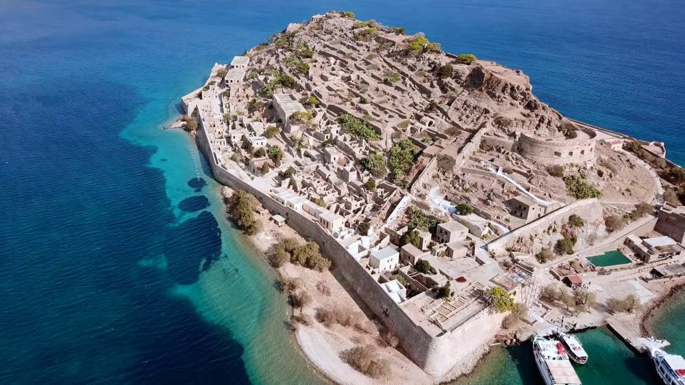 Una vista aerea dettagliata dell'isola di Spinalonga, Creta, con antiche rovine e fortificazioni circondate da acque limpide. | Noleggio auto economico