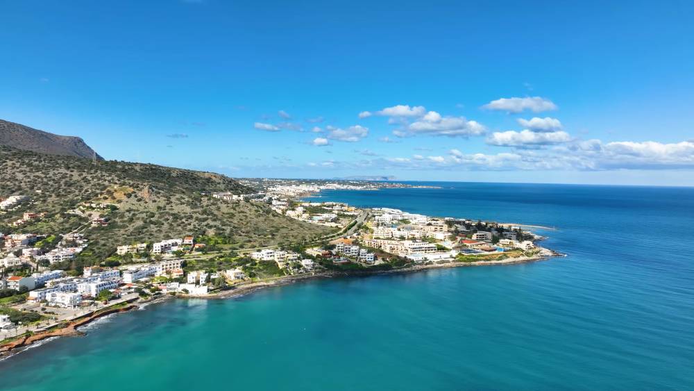 Vue aérienne de Stalis, en Crète, avec le paysage côtier, des eaux turquoise, des bâtiments résidentiels et une colline pittoresque sous un ciel bleu clair. | Smart Car Rental